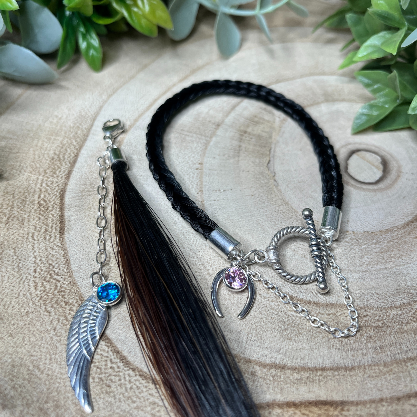 Bracelet with black hair tassel, silver charms, and gemstones on a wooden surface with greenery.