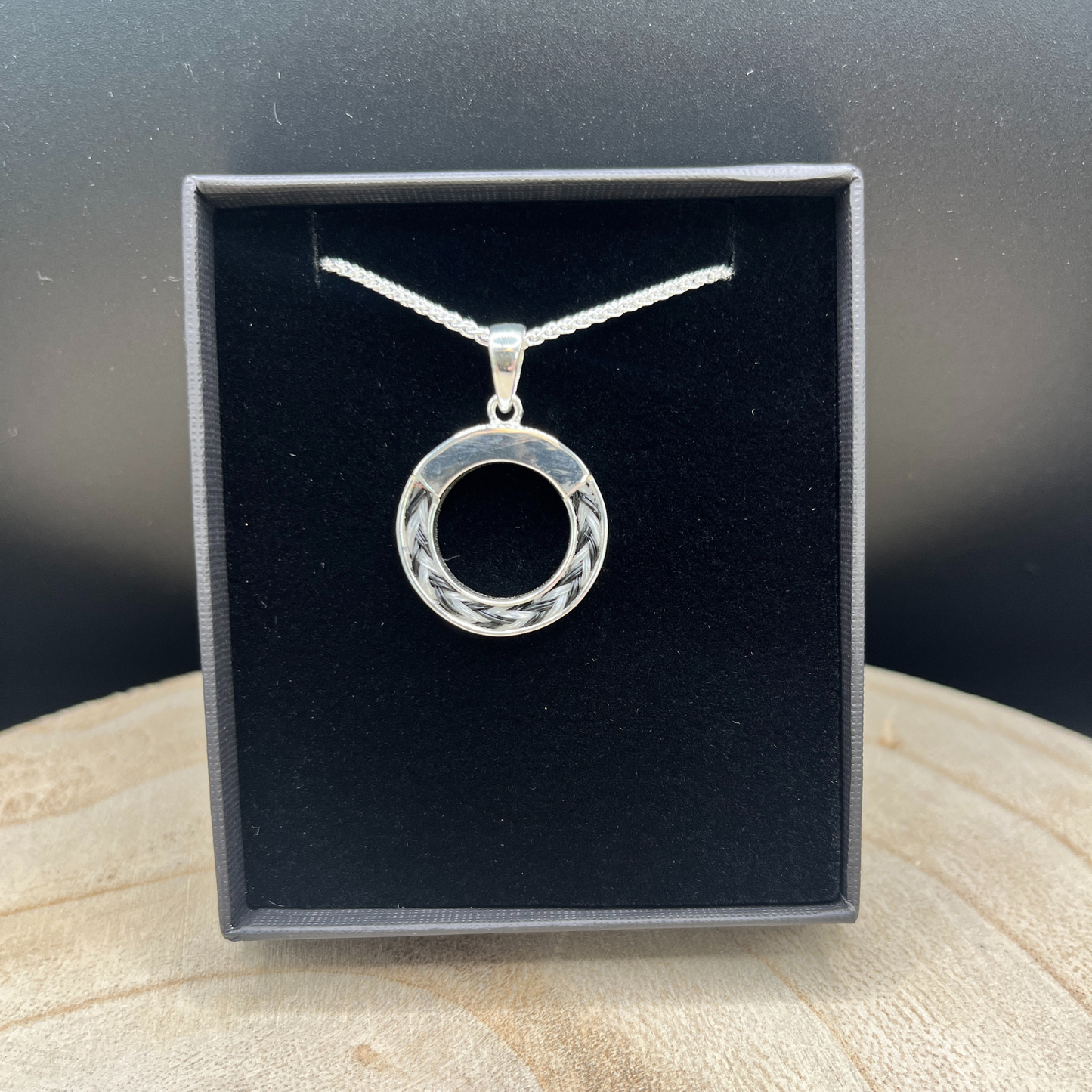 Silver necklace with a circular pendant in a black display box on a wooden surface.