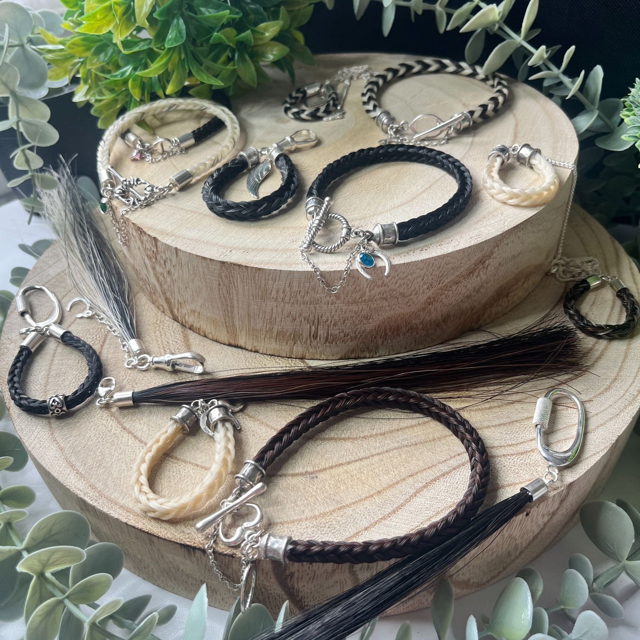Collection of braided horse hair keepsakes with charms on a wooden stand, surrounded by greenery.