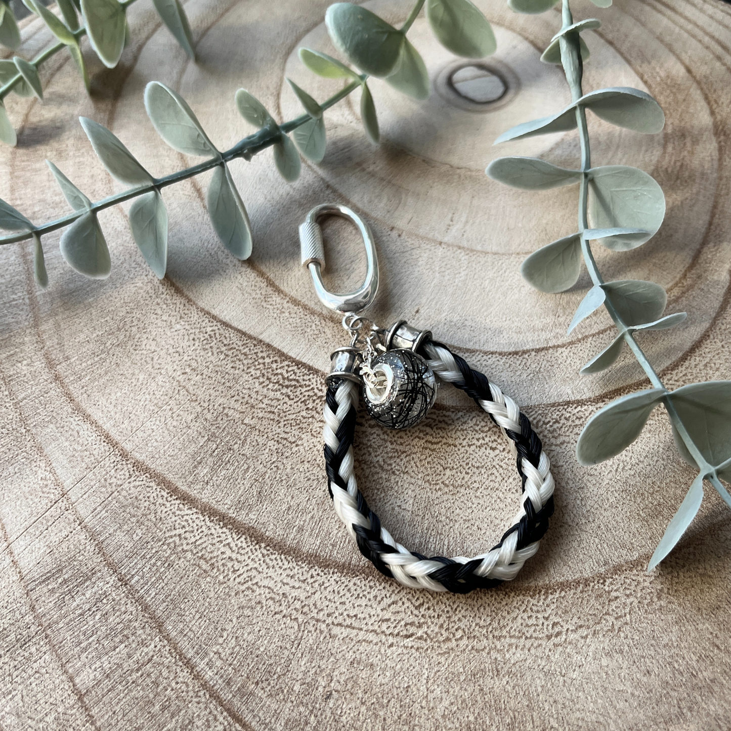 Black and white braided bracelet with a beaded charm on a wooden surface with eucalyptus leaves.